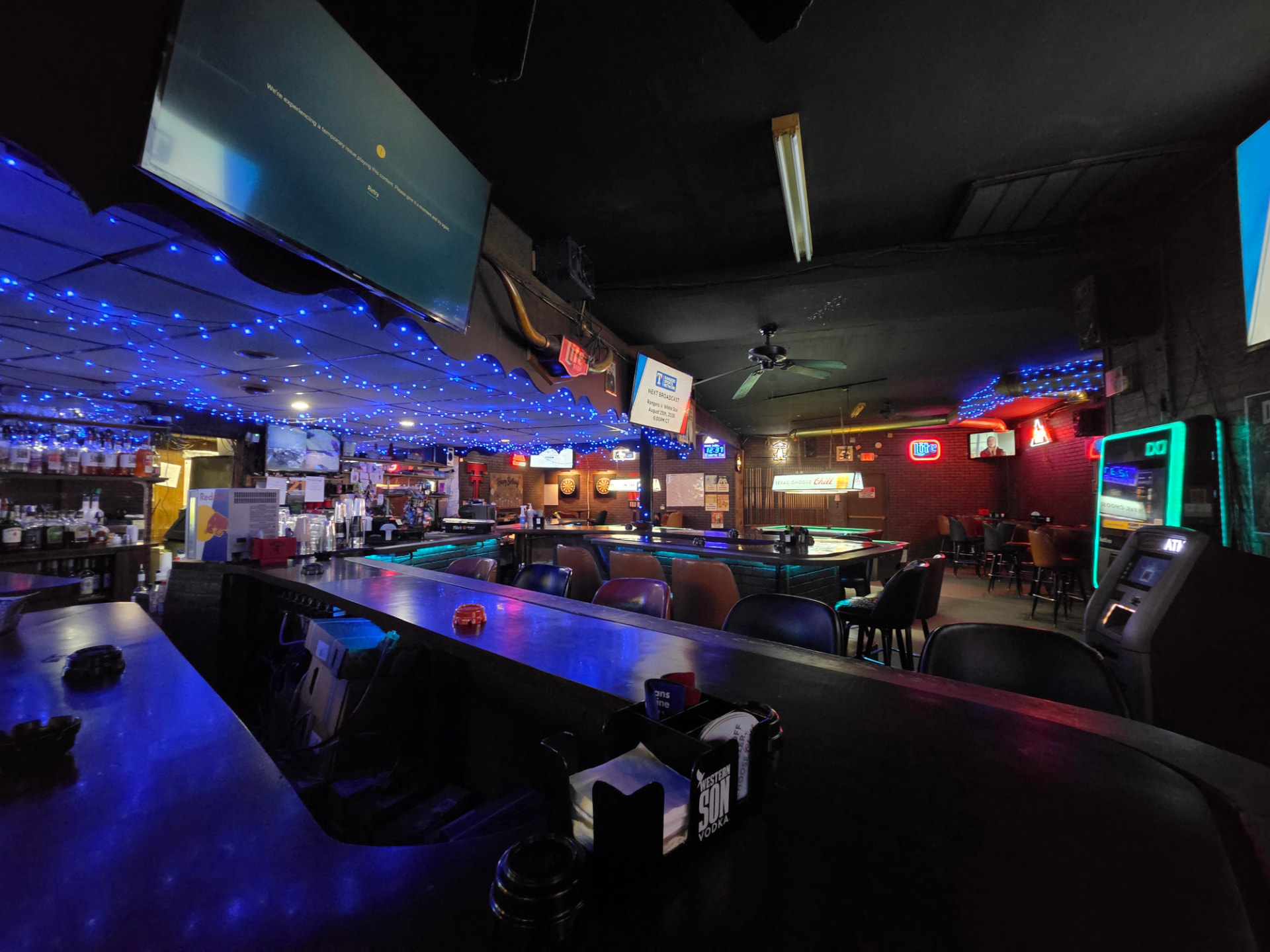Down the length of the bar under the blue string-light canopy, pool table and neon signs beyond